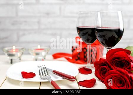 Valentines day dinner concept. Two glasses of wine, red roses, gift box, plate and burning candles on a white wooden table. Selective focus. Stock Photo