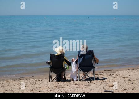 A mature, probably retired couple, gazing the Lake Huron while sitting on their folding chair at the beach. Stock Photo