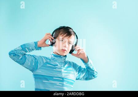 Kid enjoying music on his headphones, listening to music. Handsome young stylish kid in headphones standing against blue background Stock Photo