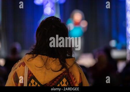 Rear view of woman wearing warm hoodie standing in auditorium watching live defocus performer performing on stage in event festival Stock Photo