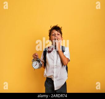 Young child student with ringing alarm clock is lazy going to school. Yellow background. Stock Photo