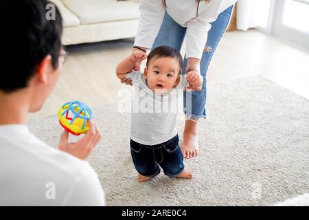 Asian baby boy toddler taking first steps. Family of father and mother encouraging their son learning to walk at home Stock Photo
