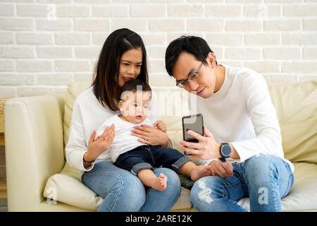 Cheerful young Asian family with son watching video on mobile phone and sitting on sofa couch at home, parents and kid enjoying cartoon movie Stock Photo