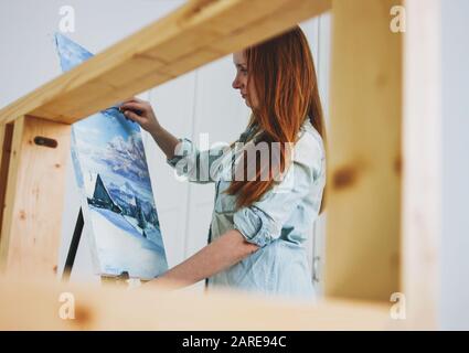 Young woman artist with long red hair draws picture with palette knife on an easel at studio Stock Photo