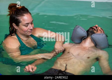 Young man with closed eyes and rubber float around neck receiving hand water massage in spa in swimming pool from female therapist  Stock Photo