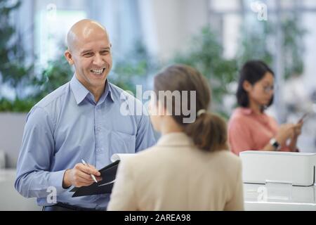 Waist up portrait of bald mature businessman smiling happily while talking to female colleague or secretary standing in modern office interior, copy s Stock Photo