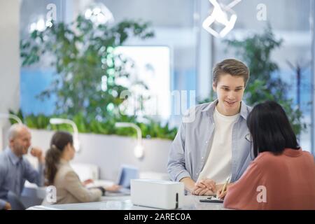 Portrait of young man talking to Asian friend while standing at tall desk, copy space Stock Photo