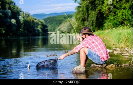 I catch it by myself. bearded man with fish on rod. hipster fishing with spoon-bait. fly fish hobby. Hipster in checkered shirt. successful fisherman in lake water. big game fishing. relax on nature. Stock Photo