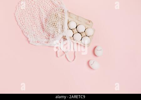 Happy Easter. White eggs with painted eyes in paper packaging and white mesh bag on pink background. Flat lay, top view, copy space. Stock Photo