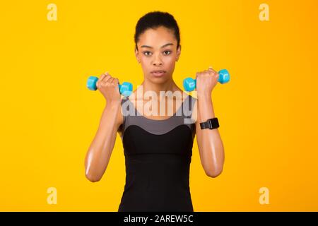 Black Girl Doing Dumbbell Bicep Curls Pumping Muscles, Yellow Background Stock Photo