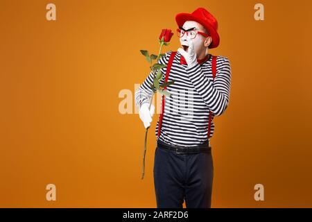 Sad mime with white face in red hat and striped t-shirt holding rose on empty orange background in studio Stock Photo