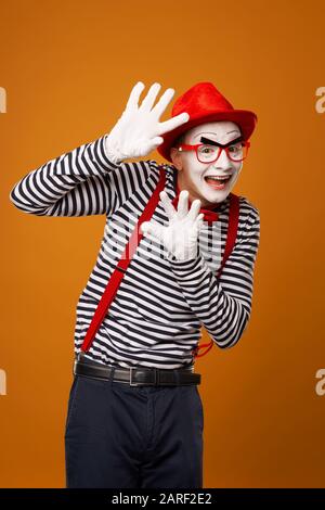Happy mime with white face in red hat and striped t-shirt on blank orange background in studio Stock Photo