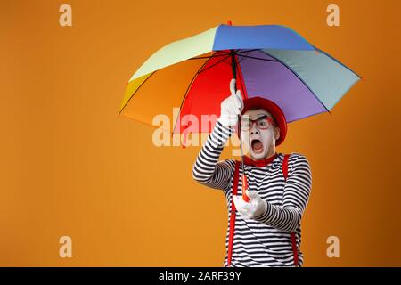 Serious mime with multi-colored umbrella on empty orange background in studio Stock Photo
