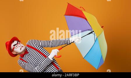 Mime with multi-colored umbrella during wind isolated on empty orange background in studio. Stock Photo