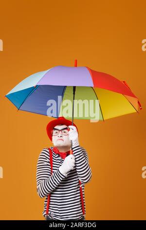 Dissatisfied mime with multi-colored umbrella isolated on blank orange background in studio Stock Photo