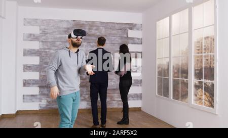 Caucasian man using virtual reality headset in empty apartment while a woman is talking with real estate agent. Stock Photo