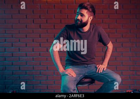 Cheerful young man in a nightclub is sitting on a bar stool near a brick wall. Neon light Stock Photo