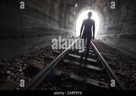Man silhouetted in a tunnel standing in the center of the railway tracks looking towards the light at the end of the tunnel in a conceptual image.