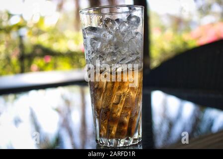 This unique photo shows an almost full glass of cola with ice. The drink is on a glass table in the garden of a restaurant in Hua Hin in Thailand Stock Photo