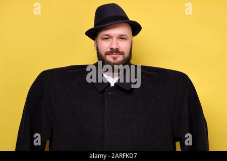 Man in oversize baggy black coat and hat smiling standing on yellow studio wall. Concept of diet and huge oversize clothes. Stock Photo