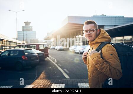 Smiling young man with backpack on car park aganist airport terminal building at sunset. Stock Photo