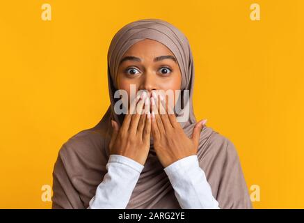 Shocked afro muslim woman in hijab covering her mouth with hands Stock Photo