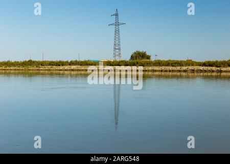 power line pylon stands on the banks of the Syr Darya river, reflected in the water, Kazakhstan. Power line crosses the river Stock Photo