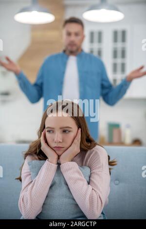 Dad standing behind a sad daughter sitting on sofa. Stock Photo