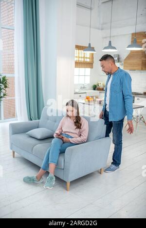 Daughter with headphones sitting on couch and father standing nearby. Stock Photo