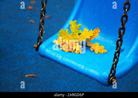 Rainy autumn day in city park. Bright blue park swing in raindrops with yellow oak leaf, selective focus. Stock Photo