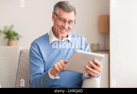 Joyful Retired Man Using Digital Tablet Sitting On Couch Indoor Stock Photo