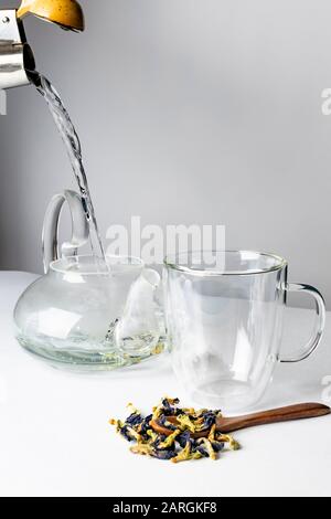 Water pours from a blue teapot into a glass mug containing dried purple ...