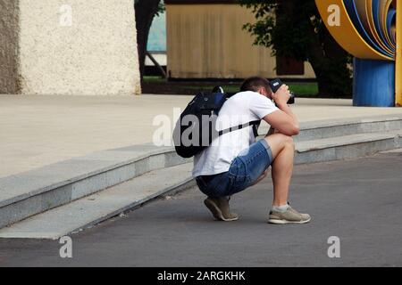 Мan wearing jeans shorts, white t-shirt, black backpack with camera and smartphone in his hands crouched down to take picture of someone or tourist at Stock Photo