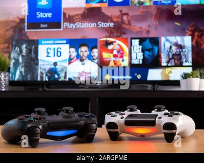 UK, Jan 2020: Pair of Sony Dualshock controllers white and black displayed in front of Playstation online store on TV