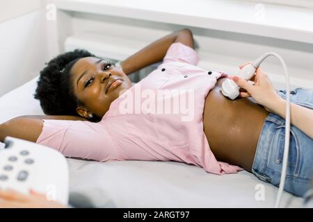 Doctor sonographer using ultrasound machine at work, holding ultrasound scanner in hand while examine abdomen of young African female patient at Stock Photo