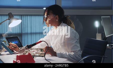 Side view of african american electronics specialist working with multimeter tester, and other electronic devices in laboratory sitting at the table in protective glasses and white robe. Stock Photo
