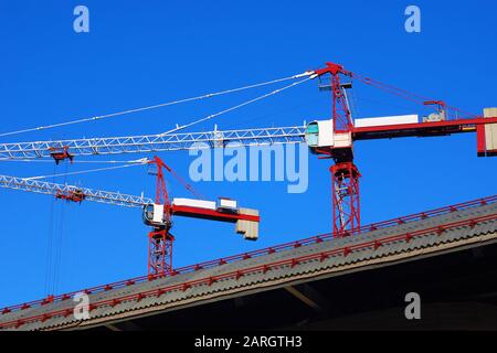 Two red and white construction cranes over a diagonal roof against blue sky Stock Photo