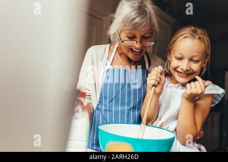 Happy grandmother and kid mixing a cake batter at home. Happy little girl stirring batter in a bowl with her granny standing by her side. Stock Photo