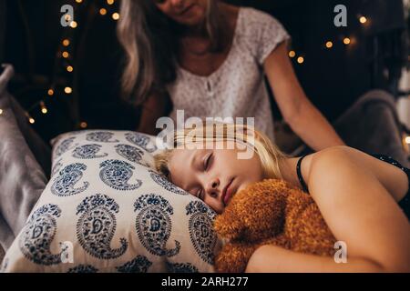Little girl sleeping holding a teddy bear. Girl sleeping on bed at home with her granny sitting by her side.