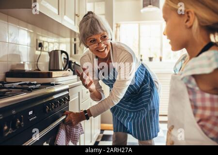 Smiling senior woman in apron opening the oven door. Happy granny and kid in kitchen cooking together. Stock Photo