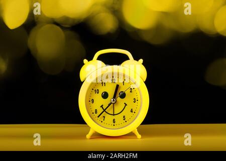 Yellow alarm clock with smililng face on the table and bokeh lights on black background. Time managing concept Stock Photo