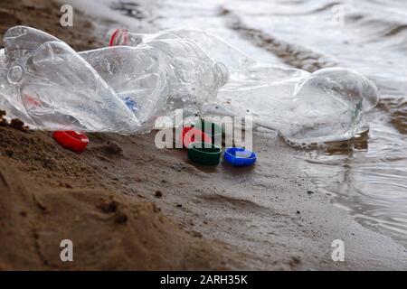 Plastic waste left by campers at river or lake shore, bottles and plastic bottle caps on the sand in the water close up Stock Photo