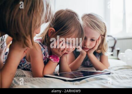 Three little sisters indoor at home using tablet lying in bed - education, technology, streaming concept Stock Photo