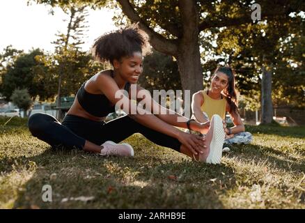 Smiling two diverse sporty fit young female friends stretching their legs in the park - african american black female warming up with her friend Stock Photo