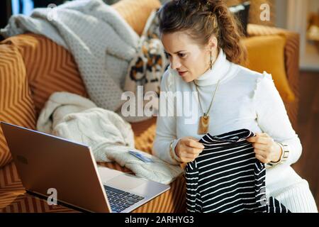 young woman in white sweater and skirt with credit card sitting near couch among old sweaters while buying new sweater shop online on a laptop at mode Stock Photo
