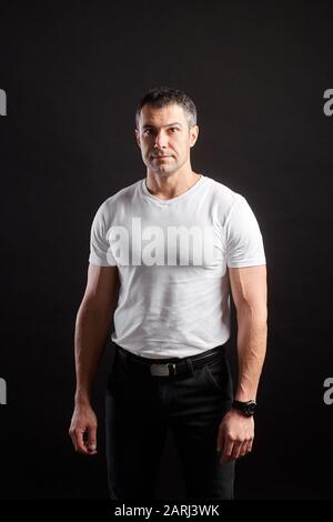 Handsome Athletic man in white blank t-shirt standing on black wall background Stock Photo