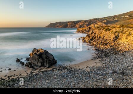 Long exposure of landmark Abano beach on the Sintra coastline in Portugal in the afternoon golden hour sun with Atlantic Ocean gentle waves lapping ag Stock Photo