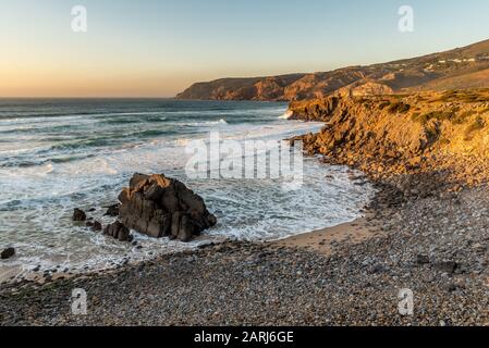 Landmark Abano beach on the Sintra coastline in Portugal in the afternoon golden hour sun with Atlantic Ocean gentle waves lapping against rocky coast Stock Photo