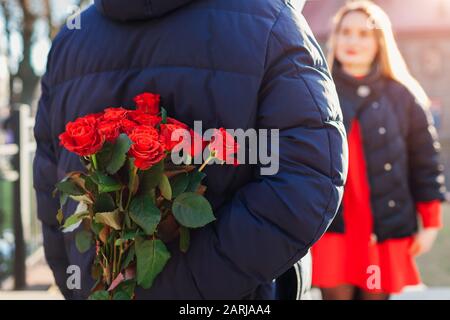 Valentines day roses. Man hiding bouquet of flowers from girlfriend behind his back on date outdoors. Womens day Stock Photo