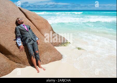 Survivor castaway businessman washed up on a tropical beach in a ragged torn suit Stock Photo